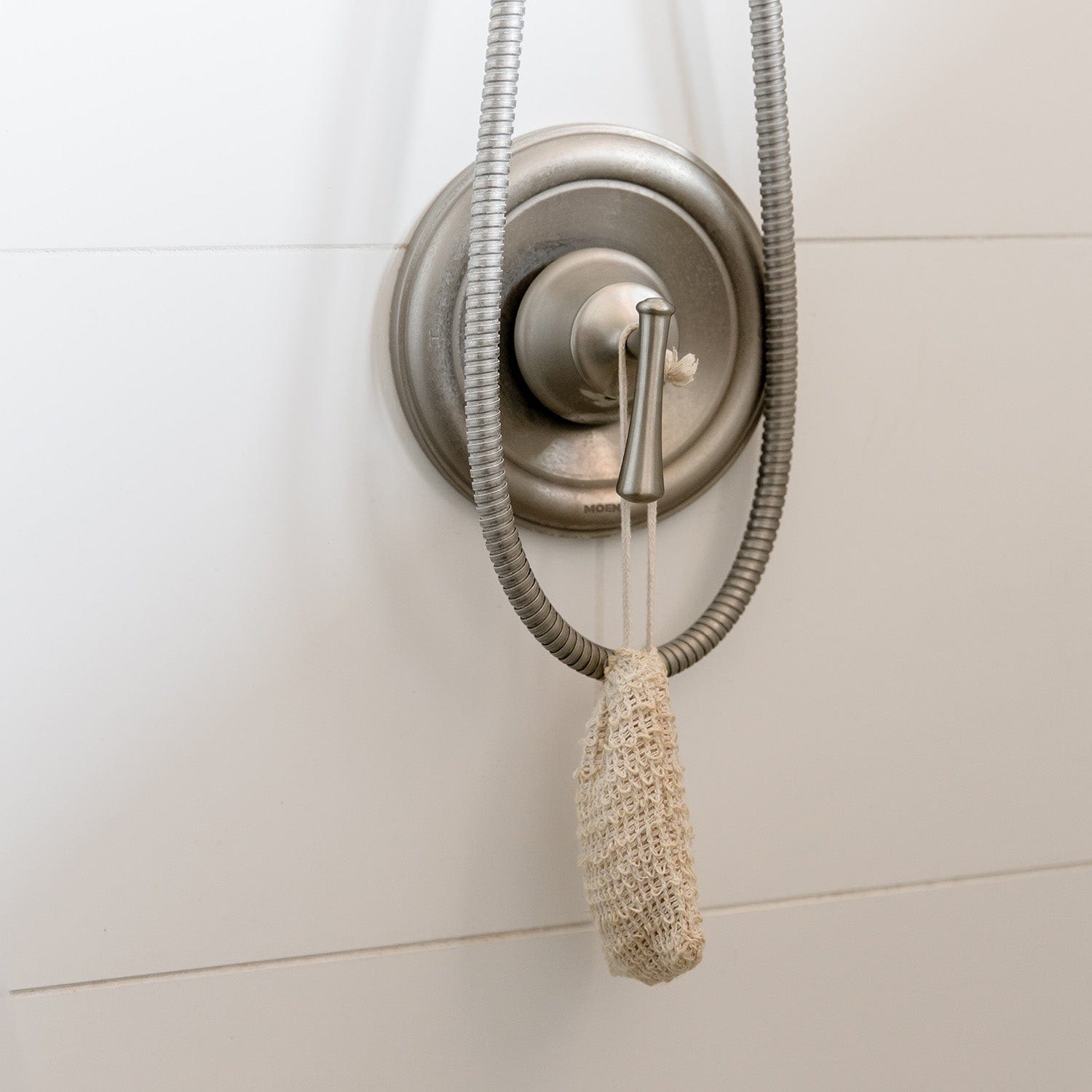 A metal shower handle with a hose is mounted on a white tiled wall. A ZeroWasteStore.com Soap Saver Bag, ideal for eco-friendly routines, hangs from the handle by its string.