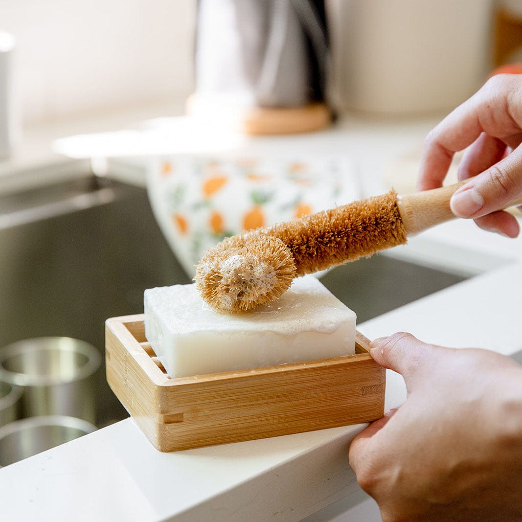 A hand uses the ZeroWasteStore.com Bottle Cleaning Brush with brown bristles, pressing it onto a white soap bar in a wooden dish by the kitchen sink.