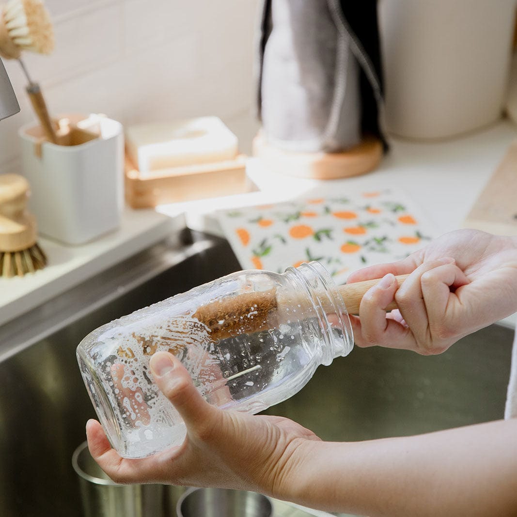 Someone uses the ZeroWasteStore.com Bottle Cleaning Brush to wash a soapy glass jar at the kitchen sink, with other cleaning items and a patterned towel visible in the background.