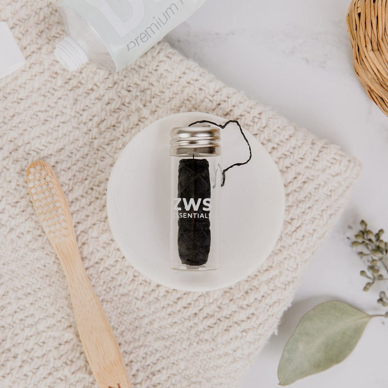 A small glass jar of Bamboo Charcoal Floss from ZeroWasteStore.com rests on a white ceramic dish beside a bamboo toothbrush, woven mat, toothpaste, and green leaves.