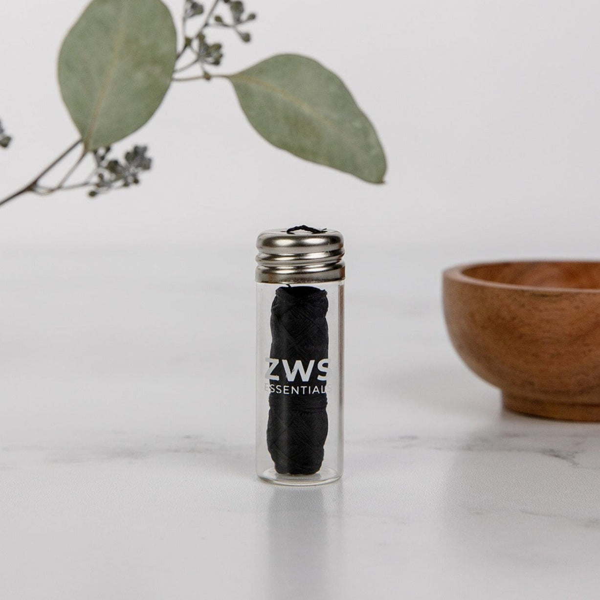 A spool of Bamboo Charcoal Floss by ZeroWasteStore.com rests in a small glass jar with a metal lid on a white surface, with a wooden bowl and eucalyptus branch behind it—your zero waste dental floss solution.