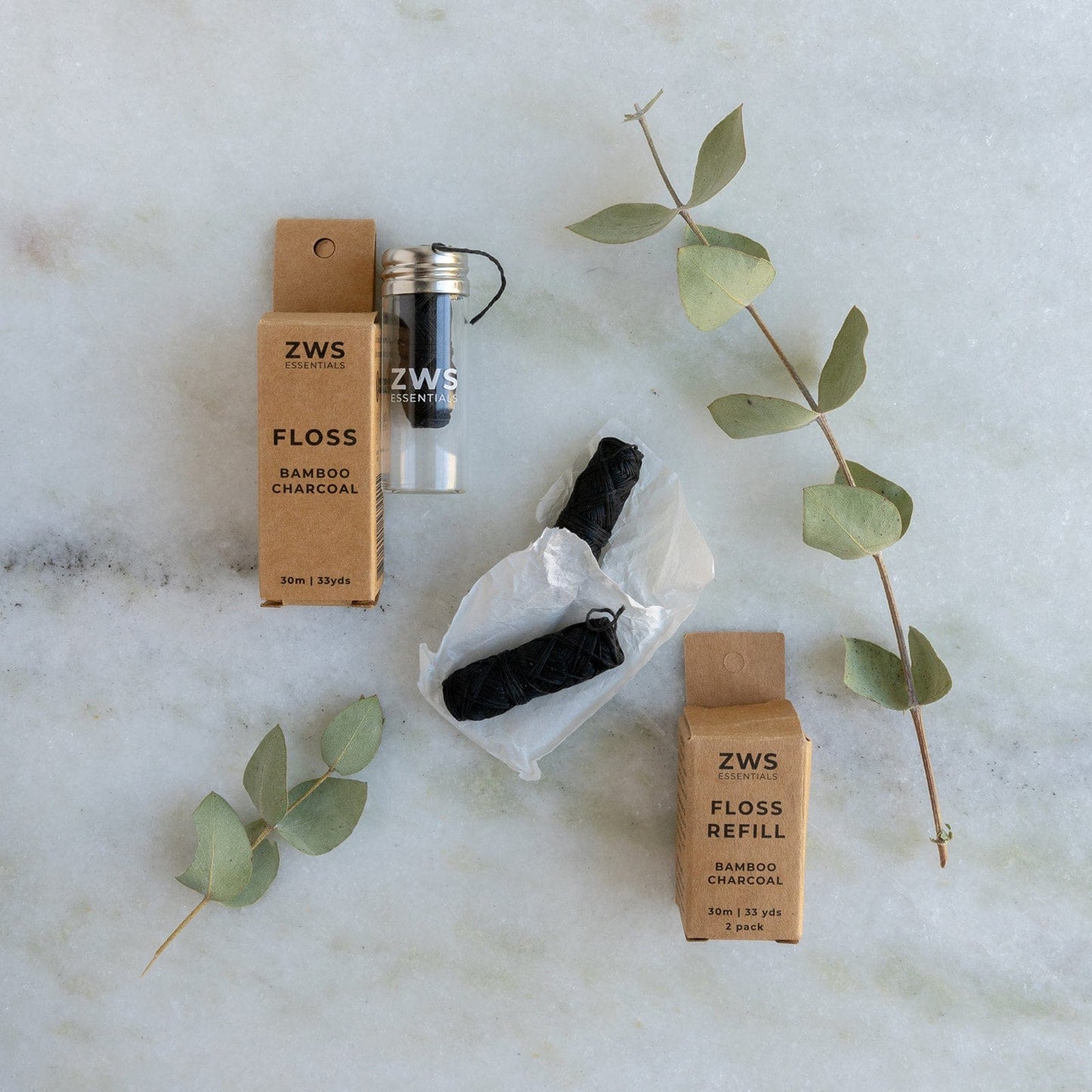 ZeroWasteStore.com Bamboo Charcoal Floss in a glass holder, with two black biodegradable refills and packaging, displayed alongside eucalyptus leaves on a light marble surface.
