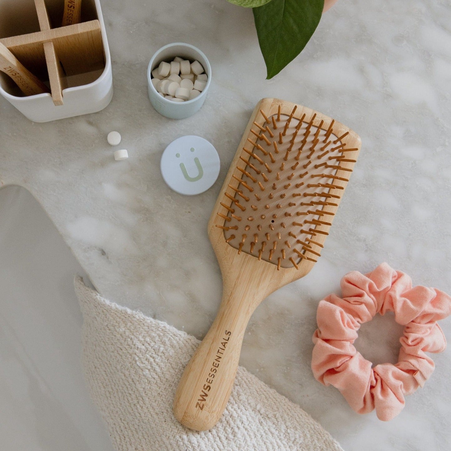 A ZeroWasteStore.com Bamboo Hair Brush sits on a marble bathroom countertop, alongside a pink scrunchie, a tablet container, and a storage box near a potted plant.