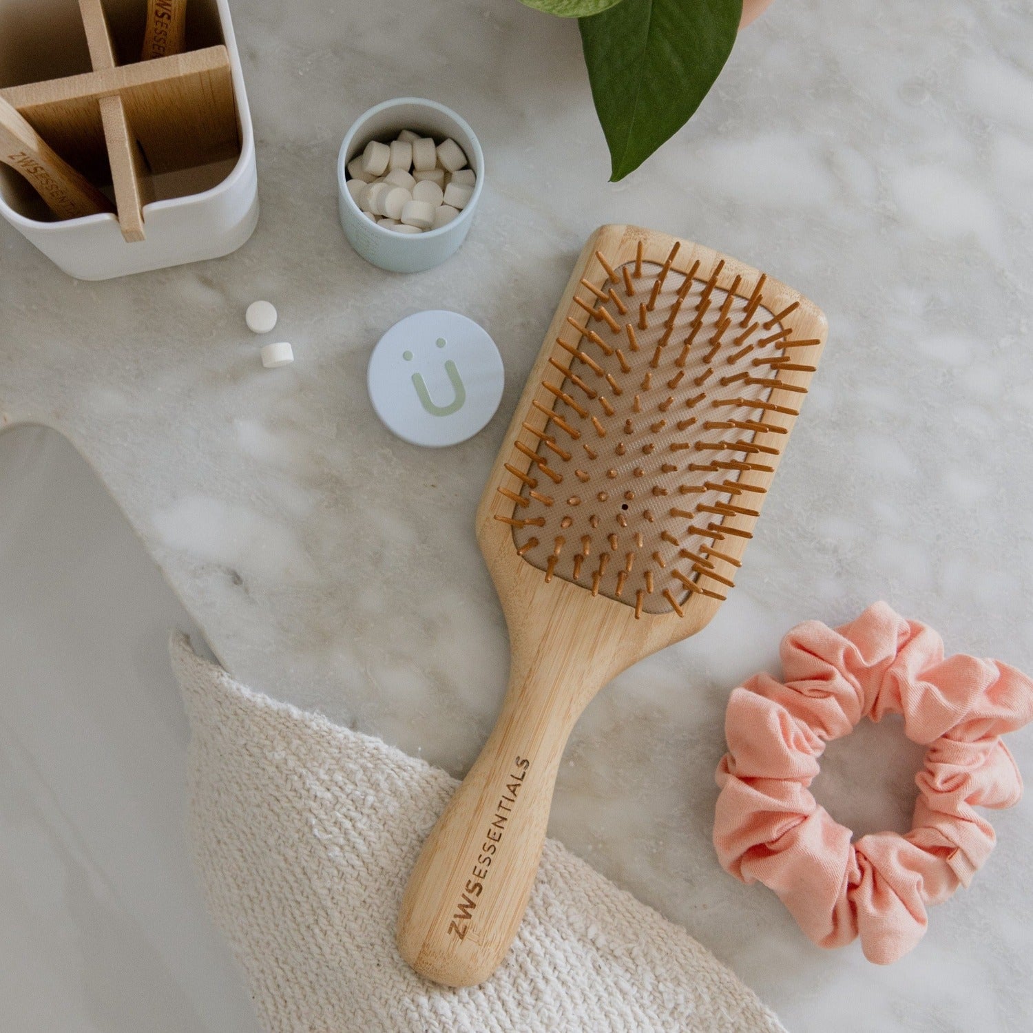 A ZeroWasteStore.com Bamboo Hair Brush sits on a marble bathroom countertop, alongside a pink scrunchie, a tablet container, and a storage box near a potted plant.