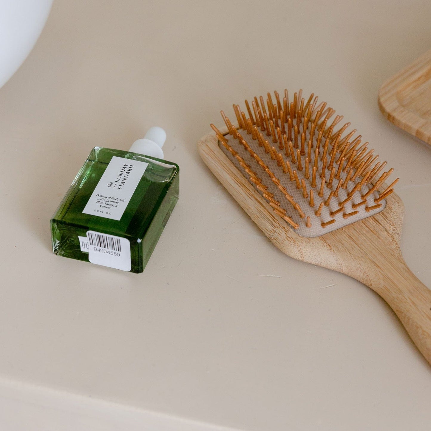 A ZeroWasteStore.com Bamboo Hair Brush with tan bristles sits beside a green glass dropper bottle labeled Scalp Serum on a beige surface.