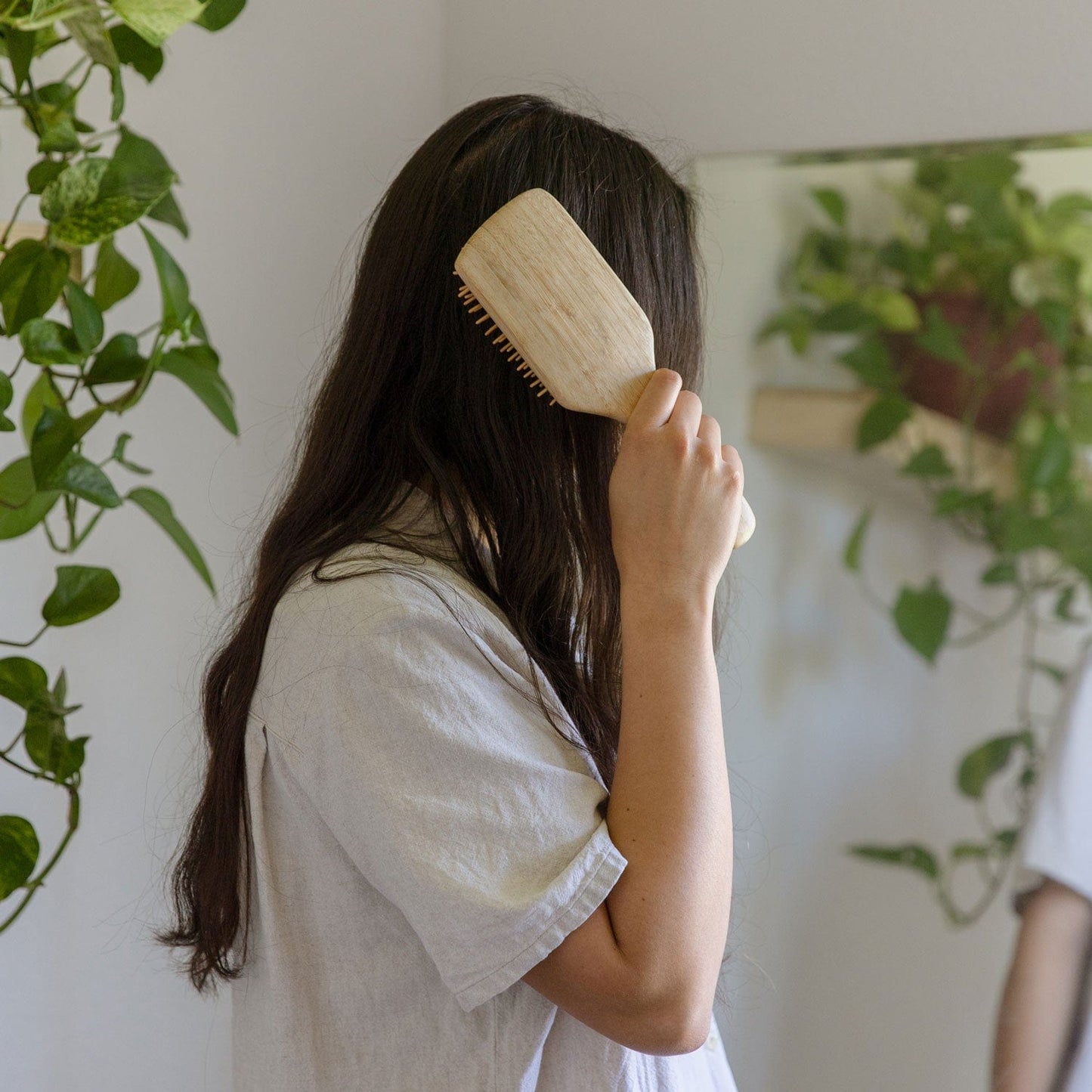 A person with long dark hair, mostly turned away, gently brushes their hair with a Bamboo Hair Brush from ZeroWasteStore.com, standing near a mirror and green plants in a light-colored shirt.
