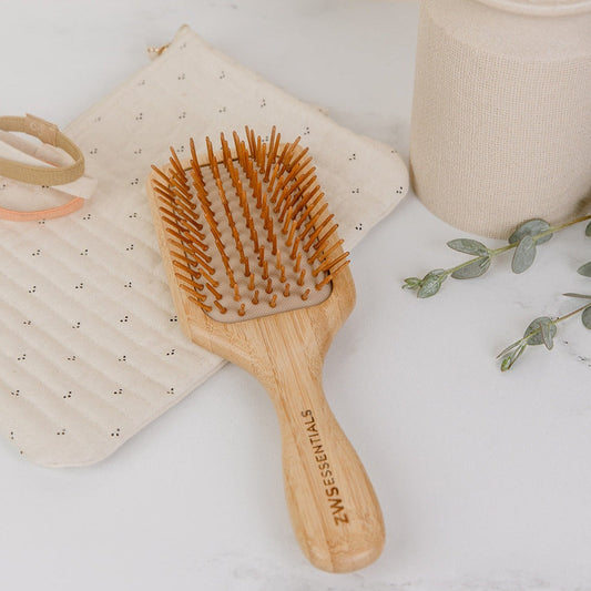 The Bamboo Hair Brush from ZeroWasteStore.com, featuring wooden bristles, sits on a quilted beige pouch beside a beige cup, light hair tie, and greenery—an ideal plastic-free choice for your eco-friendly routine.