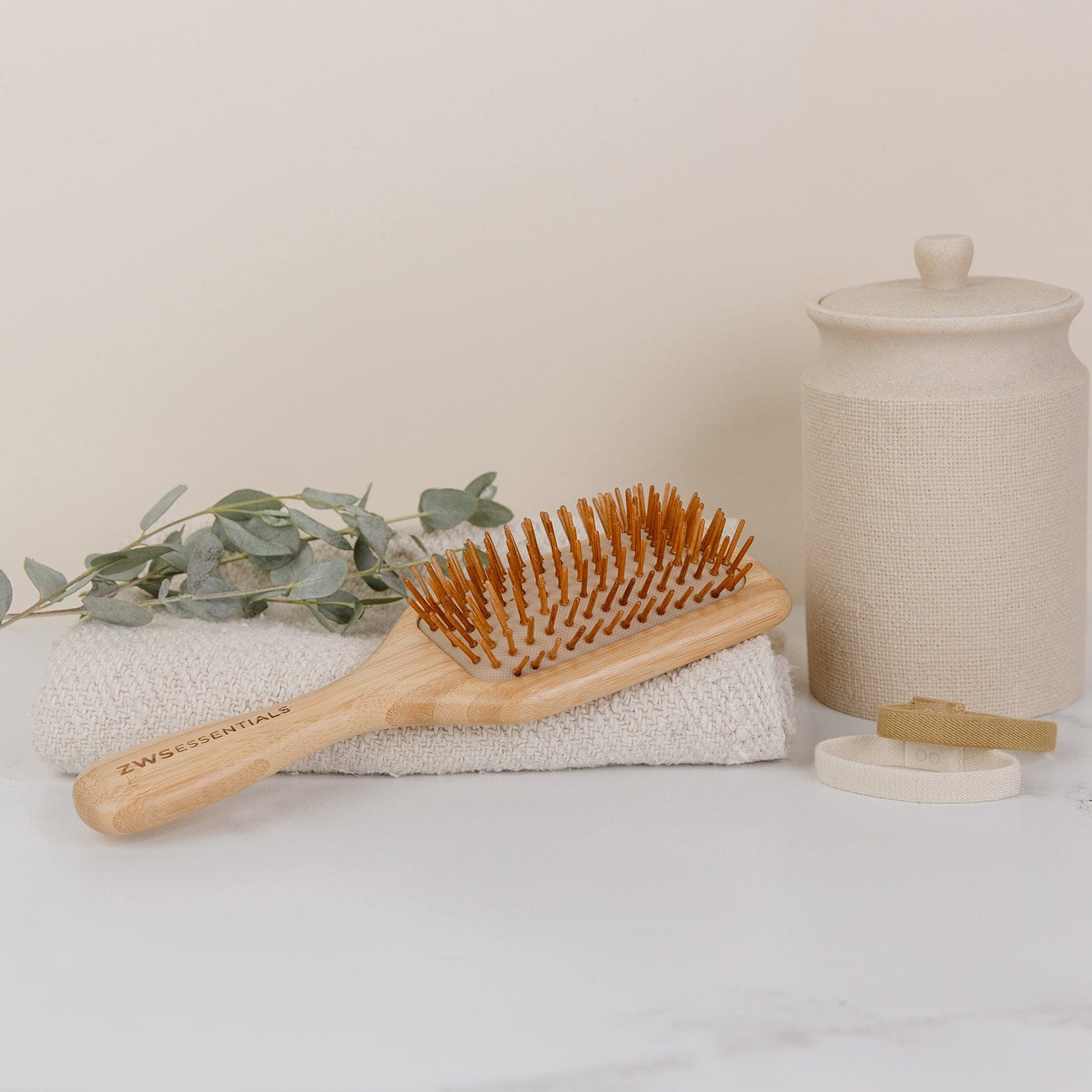 The ZeroWasteStore.com Bamboo Hair Brush is displayed on a folded towel next to green leaves, a beige lidded canister, and a small round container against a light neutral background.