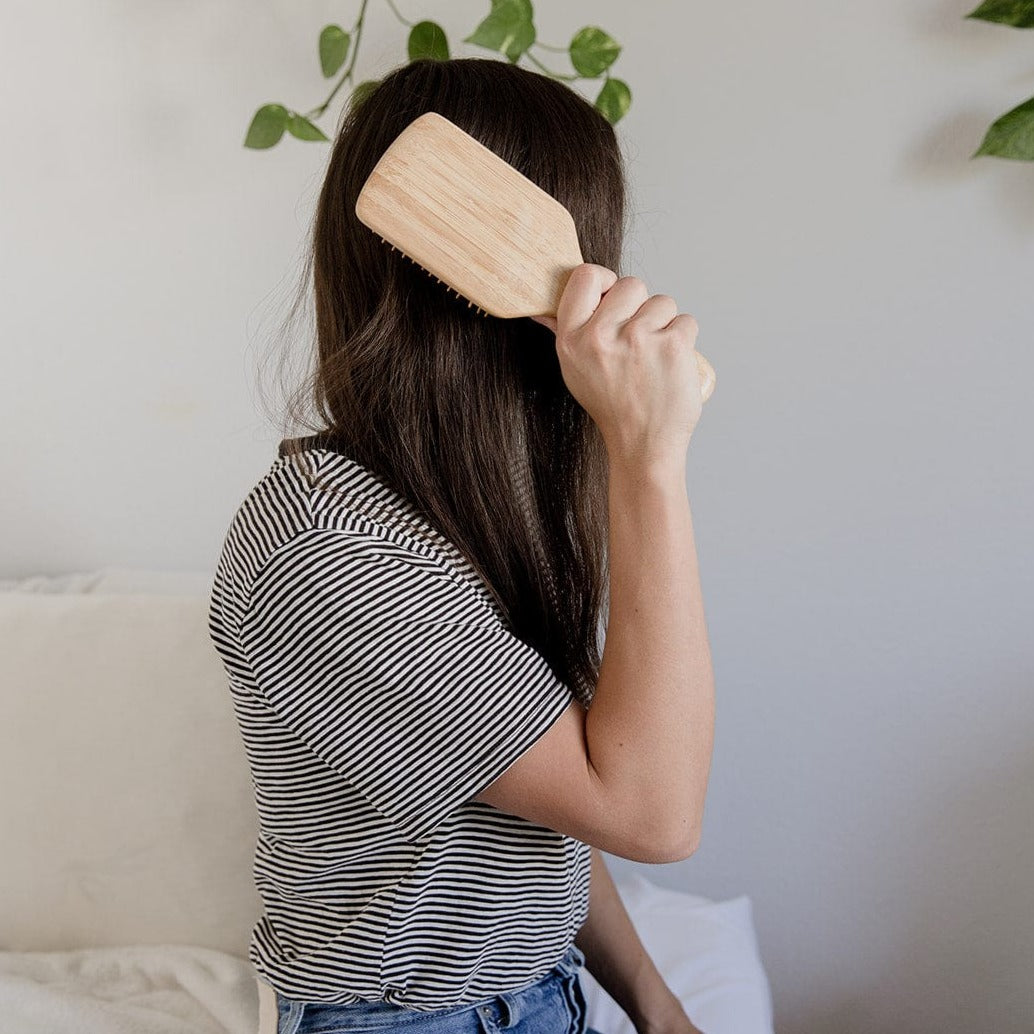 A person with long brown hair, wearing a black-and-white striped shirt, sits on a bed brushing their hair with the ZeroWasteStore.com Bamboo Hair Brush. Green leaves can be seen in the background.