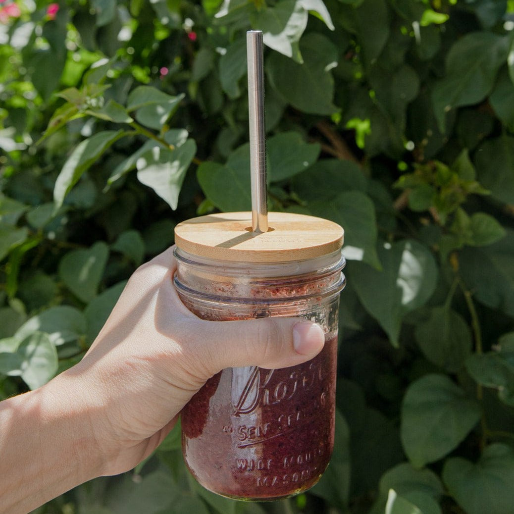 A hand holds a mason jar with a dark smoothie, topped with ZeroWasteStore.com's Bamboo Mason Jar Lid and a metal straw. Green leafy plants in the background suggest an outdoor setting.