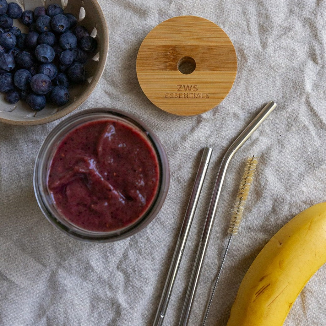 A bowl of blueberries, a smoothie in a glass jar topped with a Bamboo Mason Jar Lid by ZeroWasteStore.com, plus a cleaning brush, two metal straws, and a banana are arranged on a textured fabric surface.