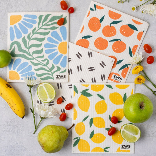 Four colorful Swedish Dish Cloths by ZeroWasteStore.com, featuring fruit and botanical patterns, are displayed on a light surface with fresh fruit and flowers—a stylish paper towel replacement for your kitchen.