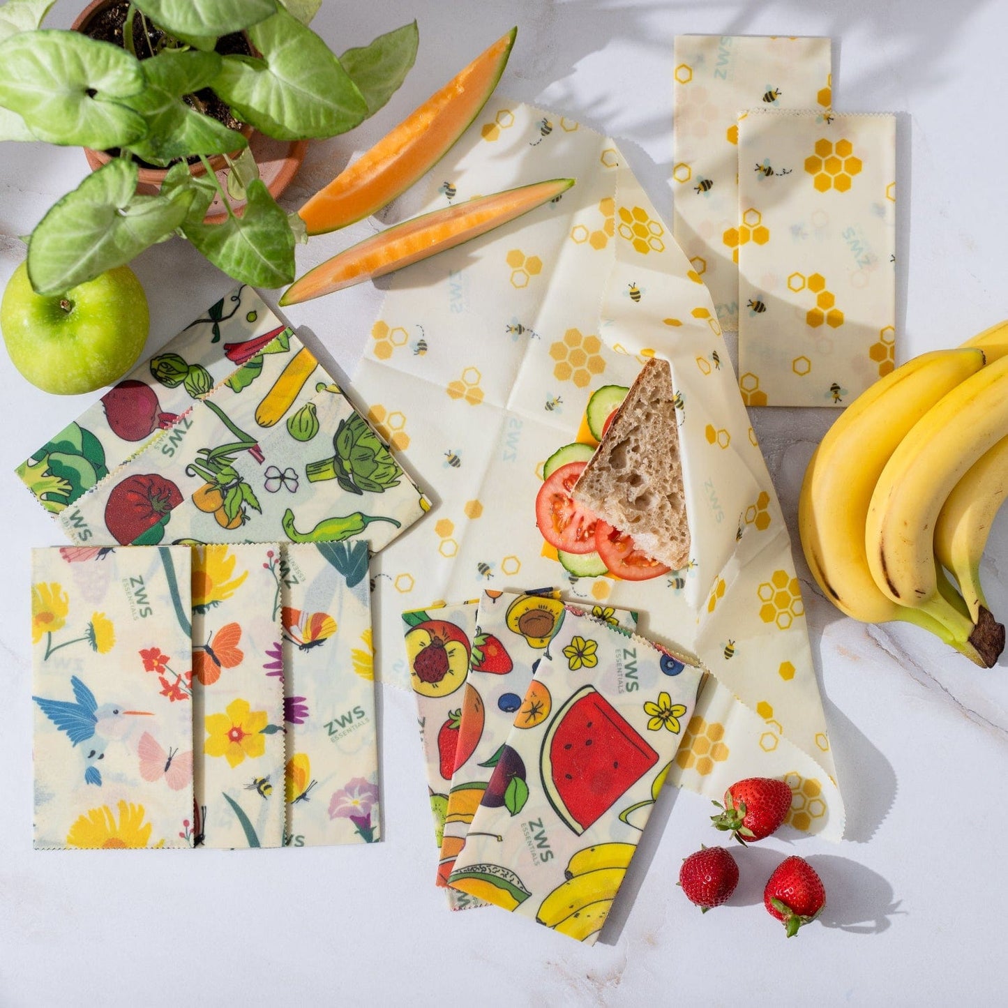 Beeswax Wraps – Plastic-Free Food Storage from ZeroWasteStore.com, featuring fruit and floral patterns, are displayed on a white surface covering bread and fruit. Fresh strawberries and a plant complete this eco-friendly alternative.