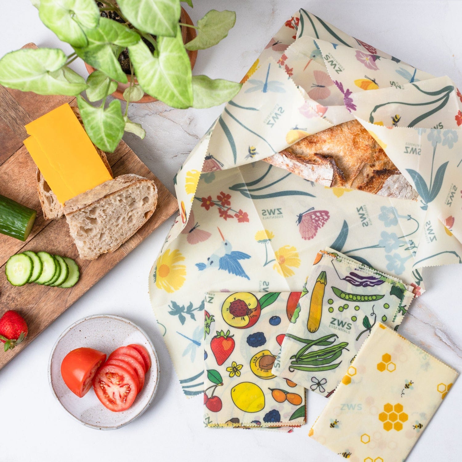 Beeswax Wraps – Plastic-Free Food Storage from ZeroWasteStore.com, featuring fruit and veggie prints, cover bread and sit beside plates of cheese, cucumber, strawberries, and tomatoes—a stylish alternative to single-use wraps.
