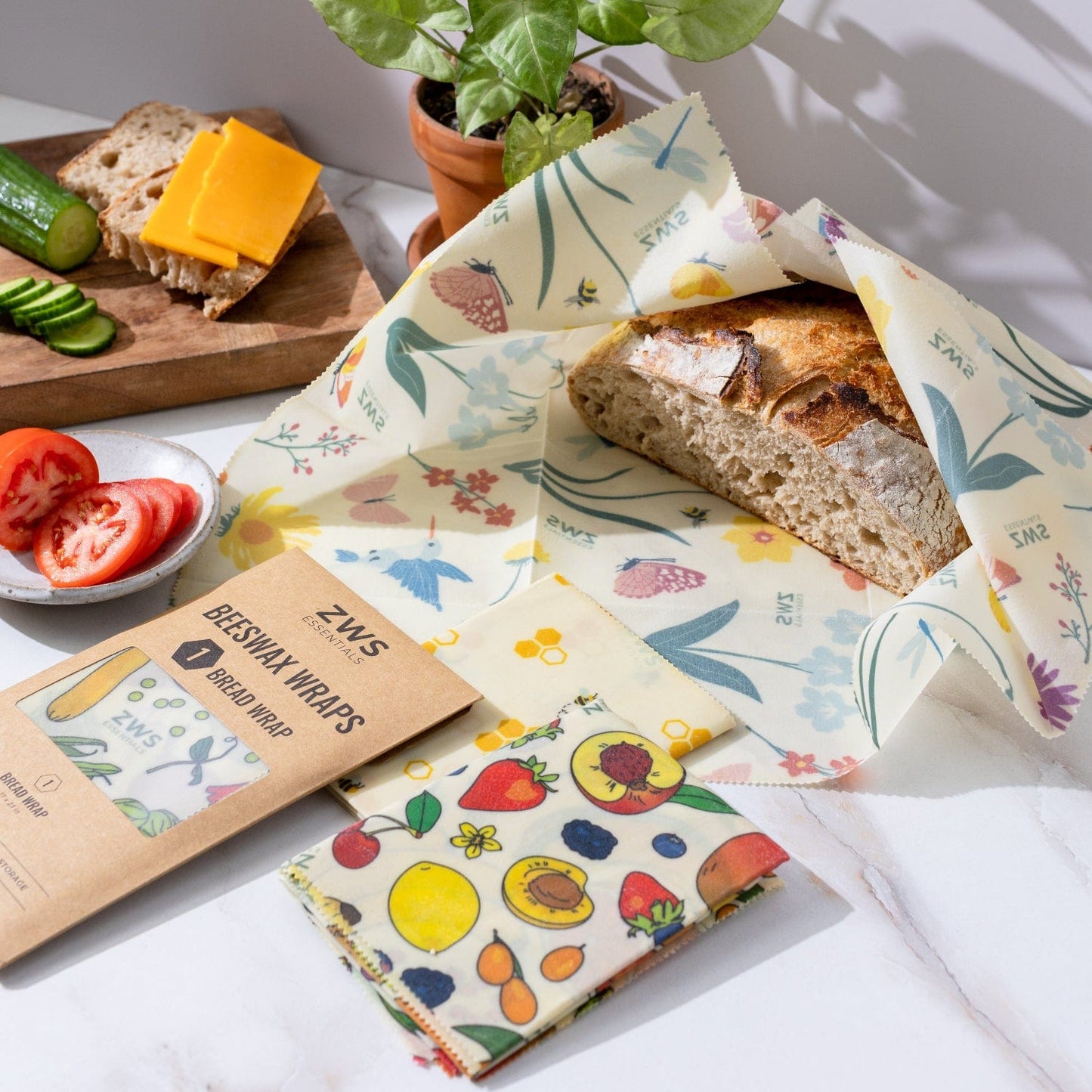 A loaf of bread wrapped in ZeroWasteStore.com’s Beeswax Wraps – Plastic-Free Food Storage sits on a marble counter near fresh veggies, cheese, a potted plant, and extra unused wraps.