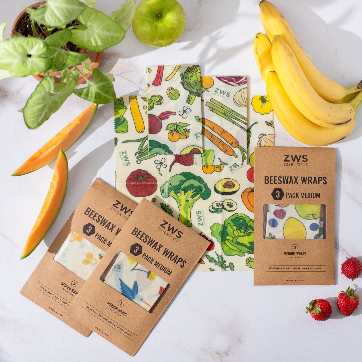 A flat lay of ZeroWasteStore.com Beeswax Wraps – Plastic-Free Food Storage with colorful veggie prints, surrounded by fruits and a potted plant. These reusable wraps come in brown packaging as an eco-friendly alternative to plastic.