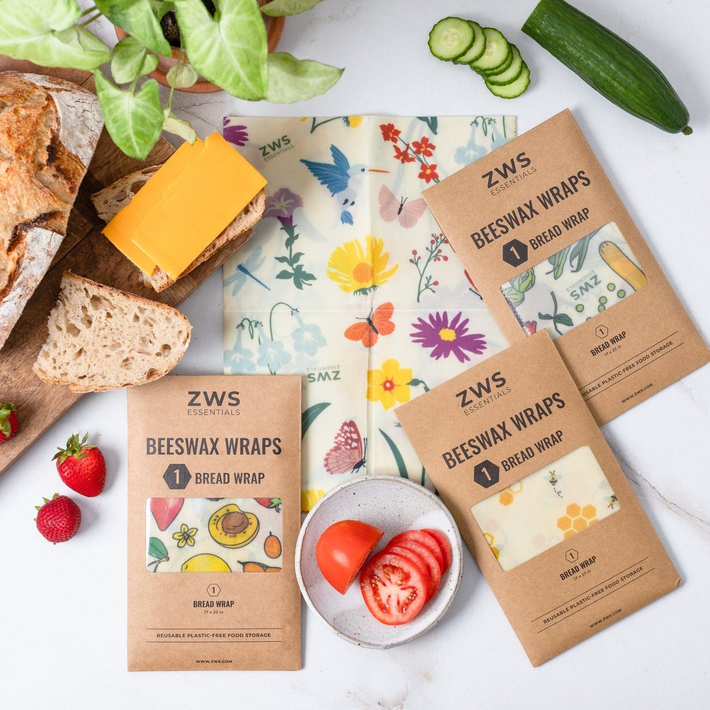 A flat lay of three ZeroWasteStore.com Beeswax Wraps – Plastic-Free Food Storage packs, one opened to show a floral wrap with bread, cheese, cucumber, strawberries, tomato slices, and a plant on a white surface.