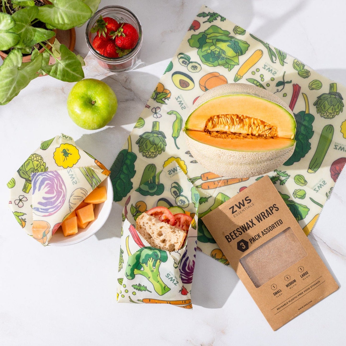 A cantaloupe, green apple, and strawberries rest on a white surface with bread and cantaloupe pieces wrapped in ZeroWasteStore.com Beeswax Wraps – Plastic-Free Food Storage. The 3-pack box is also displayed.