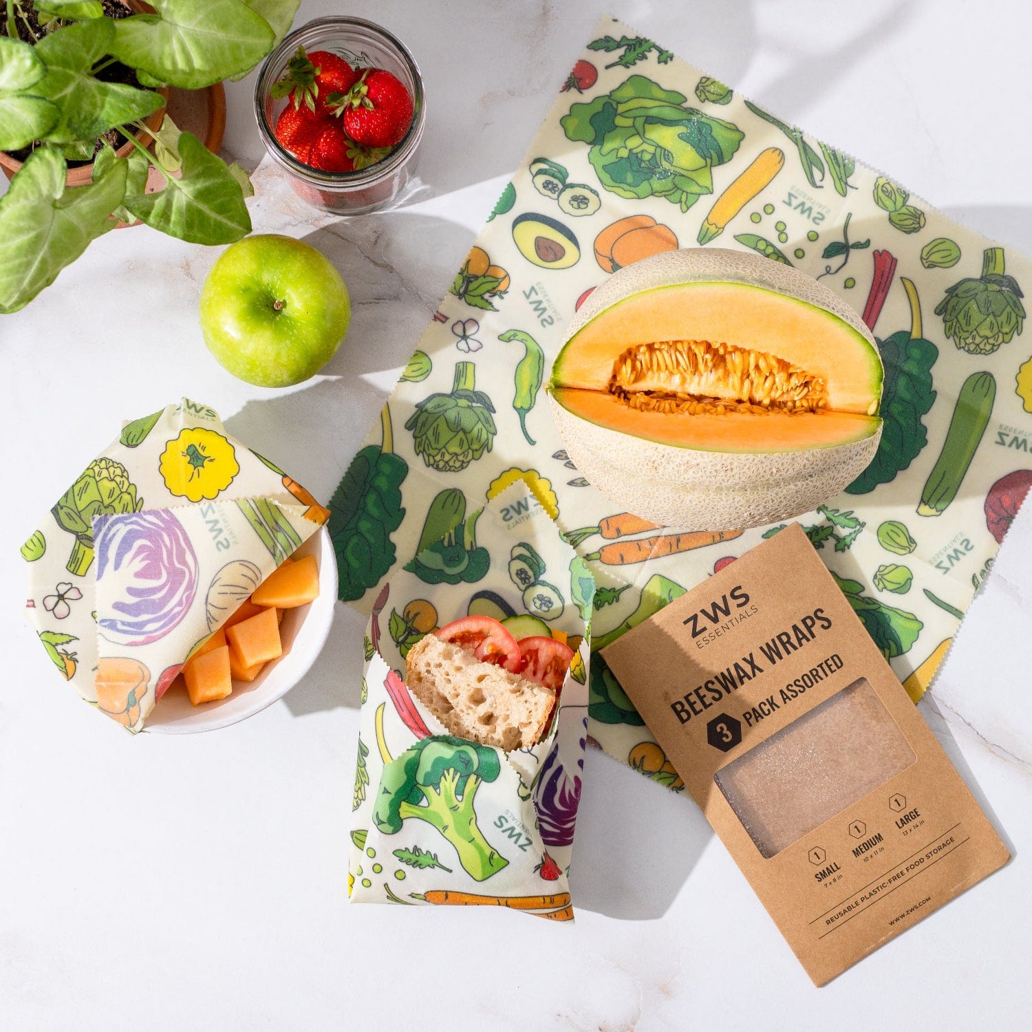 A cantaloupe, green apple, and strawberries rest on a white surface with bread and cantaloupe pieces wrapped in ZeroWasteStore.com Beeswax Wraps – Plastic-Free Food Storage. The 3-pack box is also displayed.
