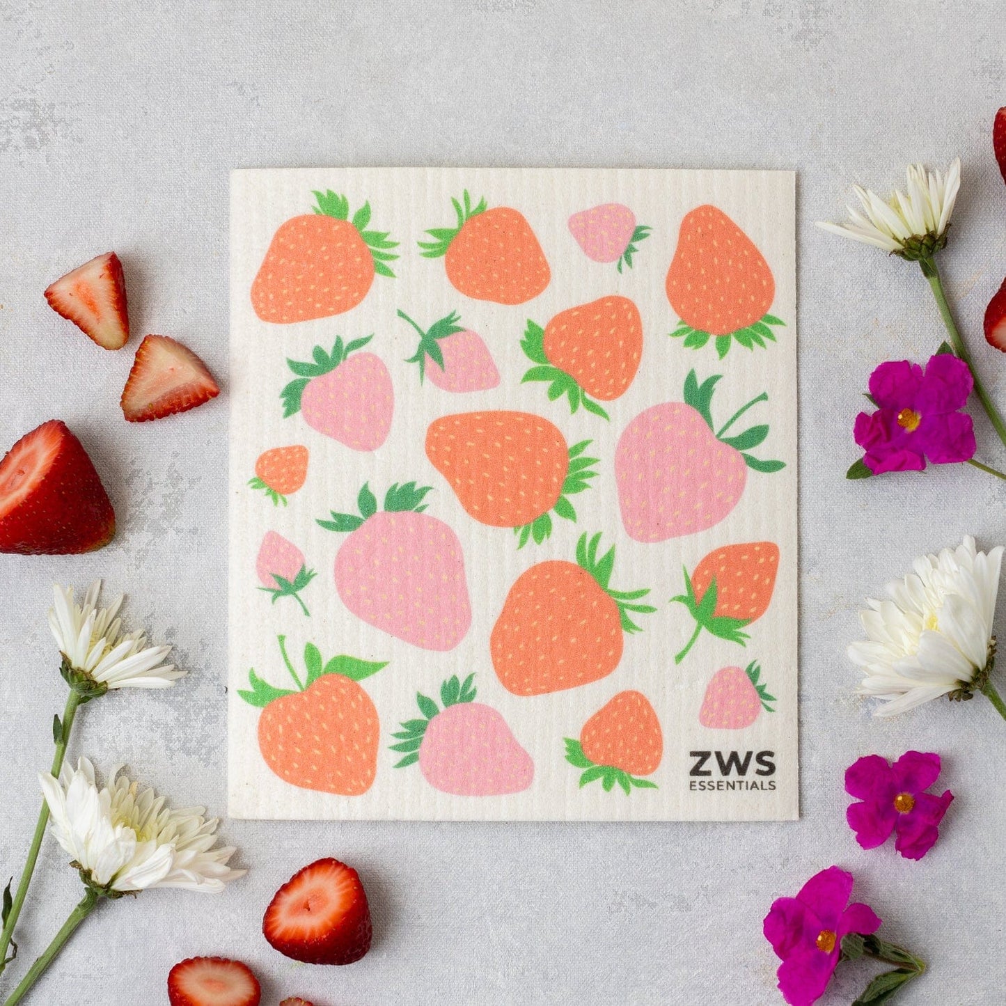 A reusable Swedish Dish Cloth by ZeroWasteStore.com with a strawberry design rests on a light surface, surrounded by fresh strawberries and flowers. The eco-friendly cloth is printed with ZeroWasteStore.com in the corner.