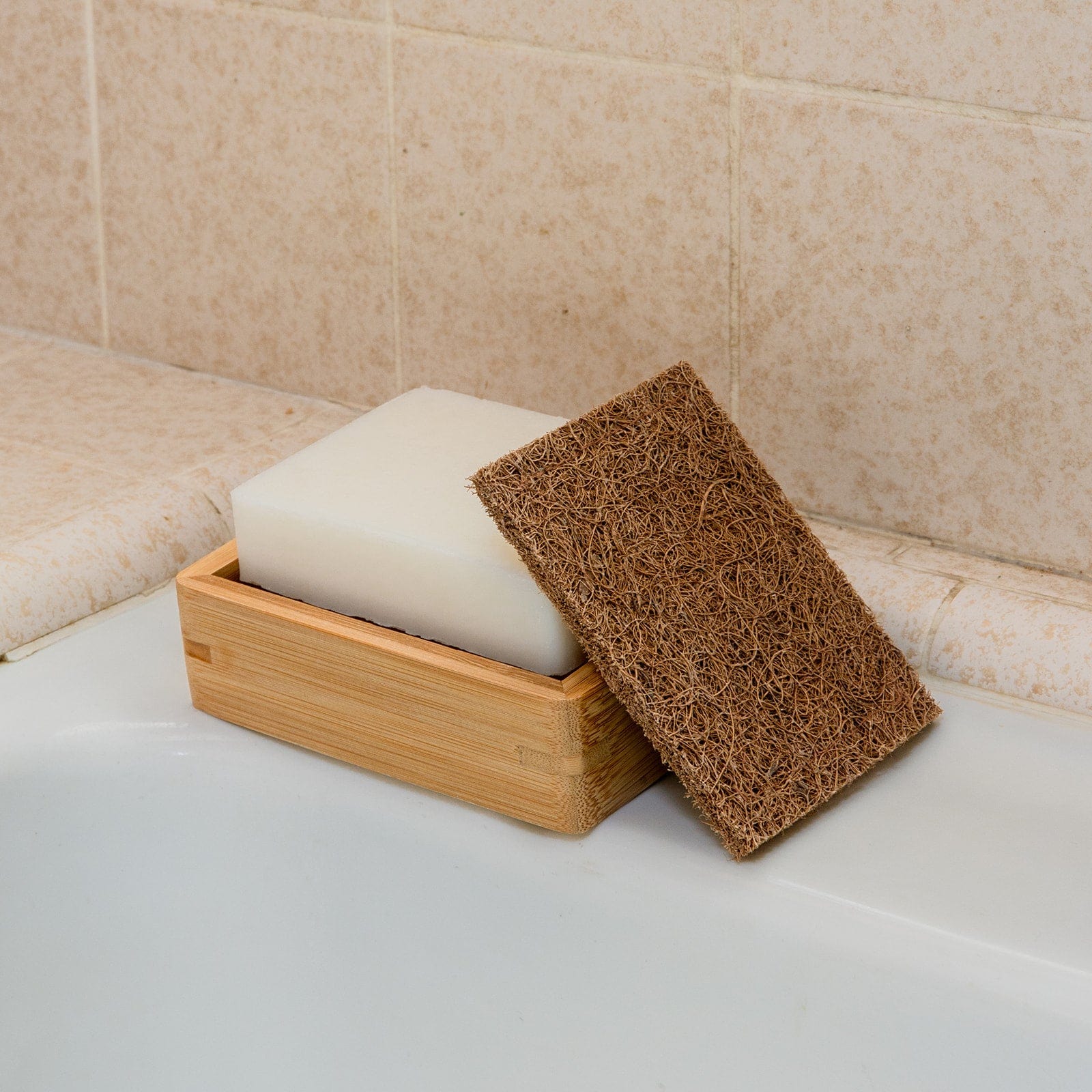 A rectangular white soap rests in a wooden dish beside the Coconut Kitchen Scrubber by ZeroWasteStore.com, placed on the edge of a bathtub with beige tiled walls.