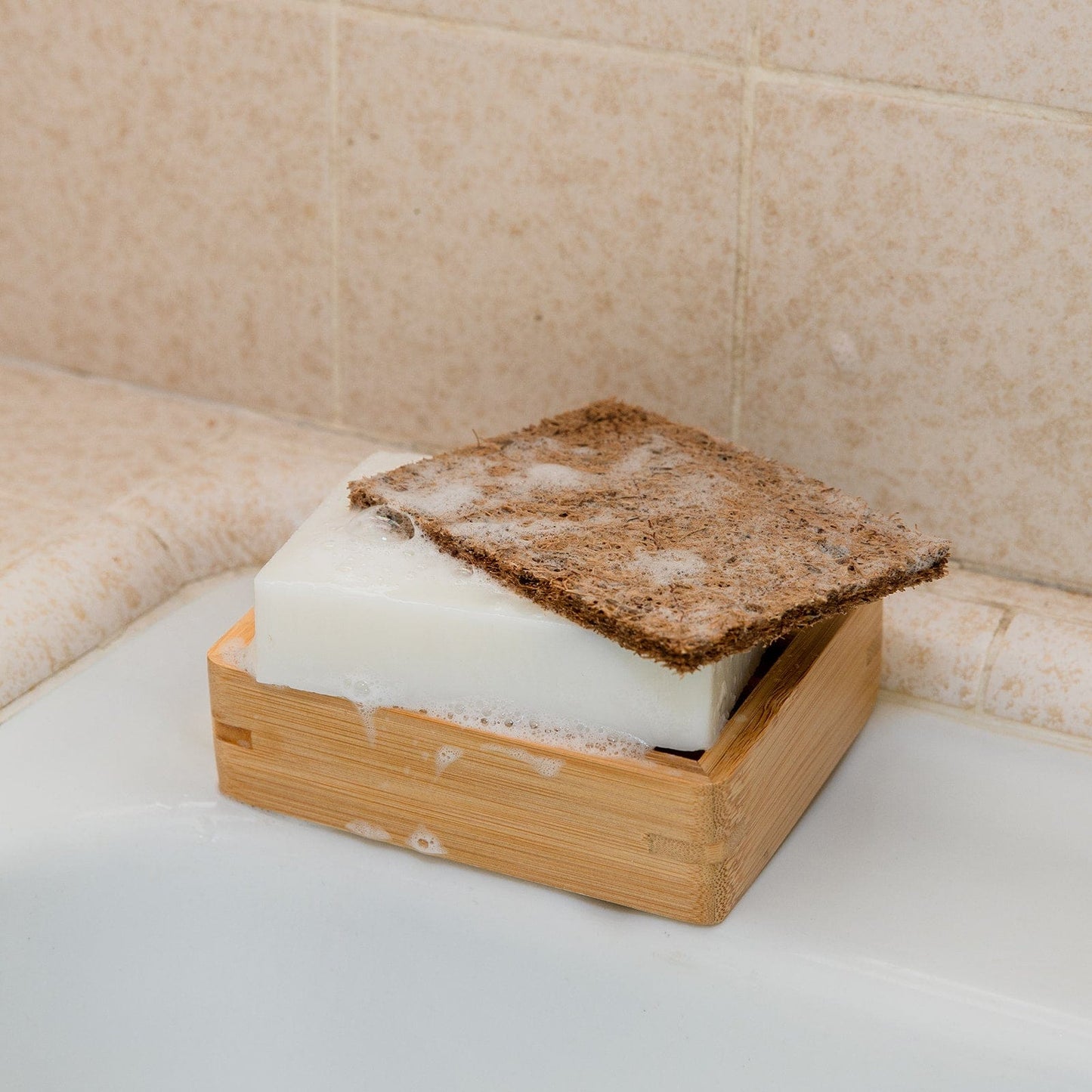 A bar of white soap with suds sits in a wooden soap dish on a bathtub edge, topped by the ZeroWasteStore.com Coconut Kitchen Scrubber, a brown rectangular biodegradable scrubber.