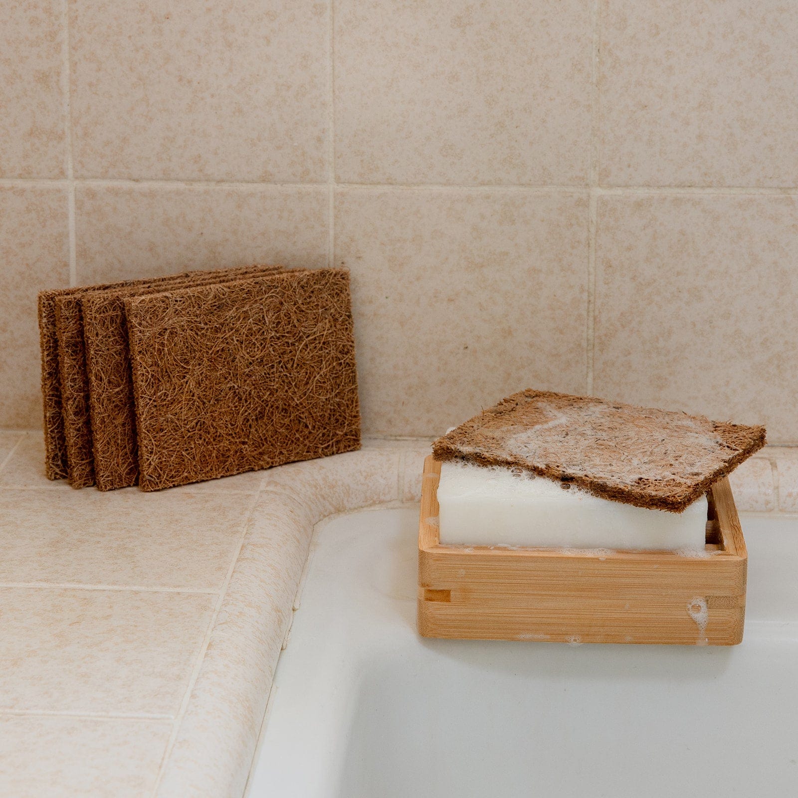 A Coconut Kitchen Scrubber by ZeroWasteStore.com rests on a white soap bar in a wooden dish by the sink, with three more brown loofah pads from the set stacked nearby on the tiled countertop.