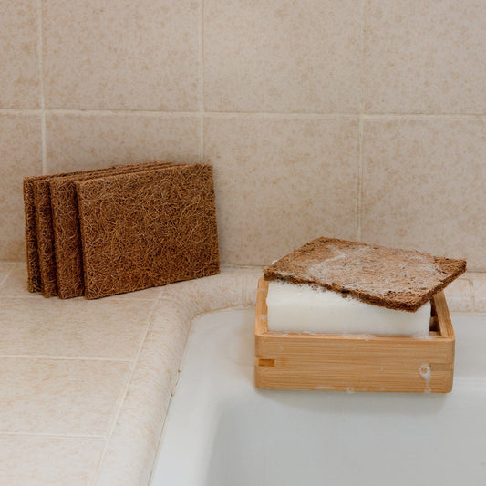 A Coconut Kitchen Scrubber by ZeroWasteStore.com rests on a white soap bar in a wooden dish by the sink, with three more brown loofah pads from the set stacked nearby on the tiled countertop.
