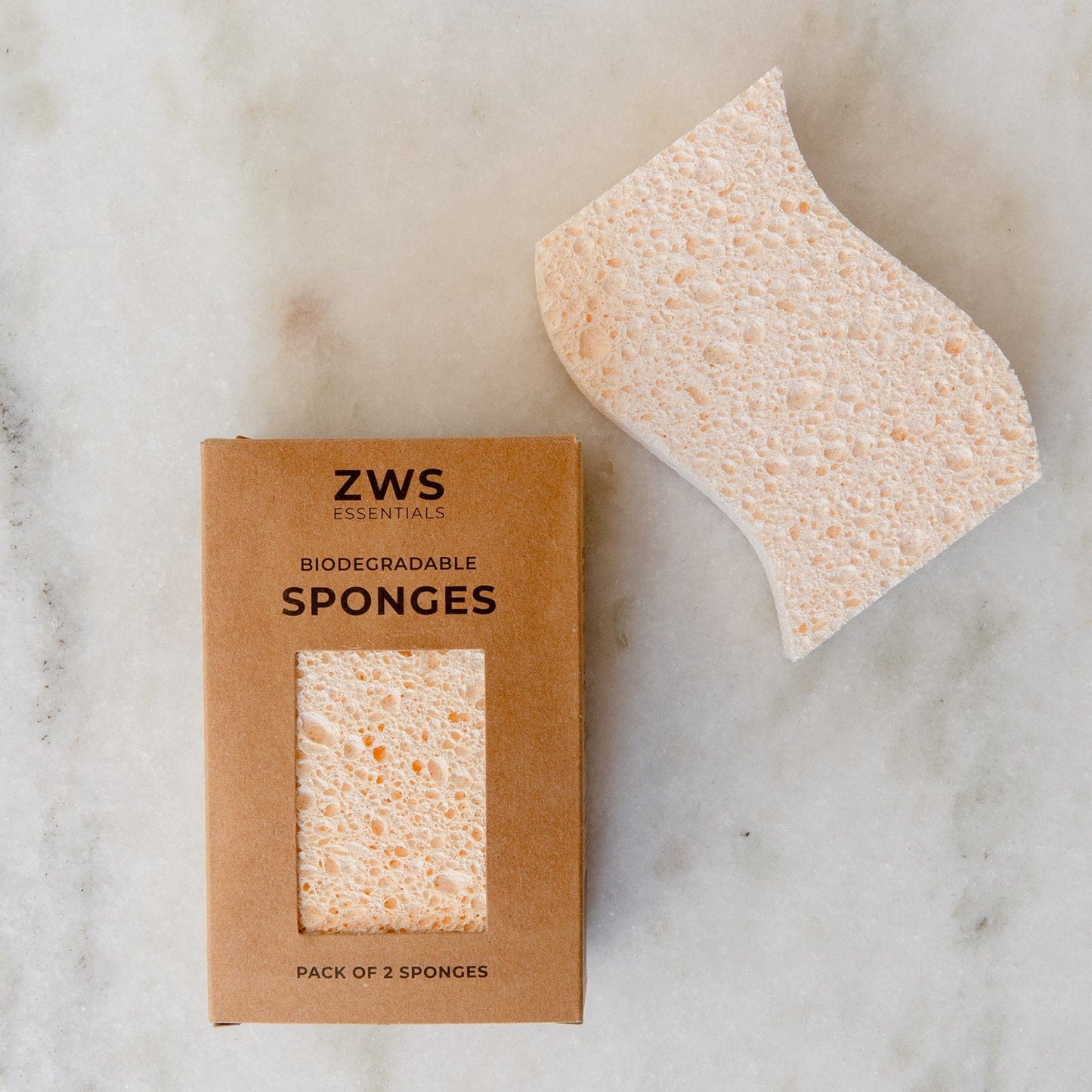 A rectangular ZeroWasteStore.com Cellulose Kitchen Sponges box sits beside a single, beige, wavy-edged sponge on a light marble surface. The box contains two biodegradable wood pulp sponges.