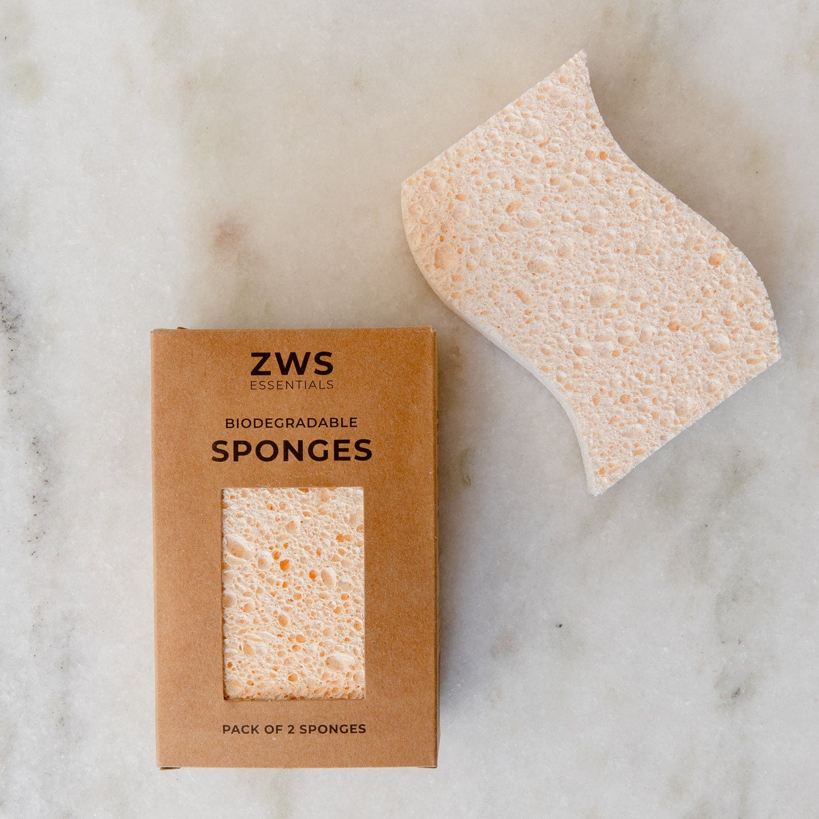 A rectangular ZeroWasteStore.com Cellulose Kitchen Sponges box sits beside a single, beige, wavy-edged sponge on a light marble surface. The box contains two biodegradable wood pulp sponges.