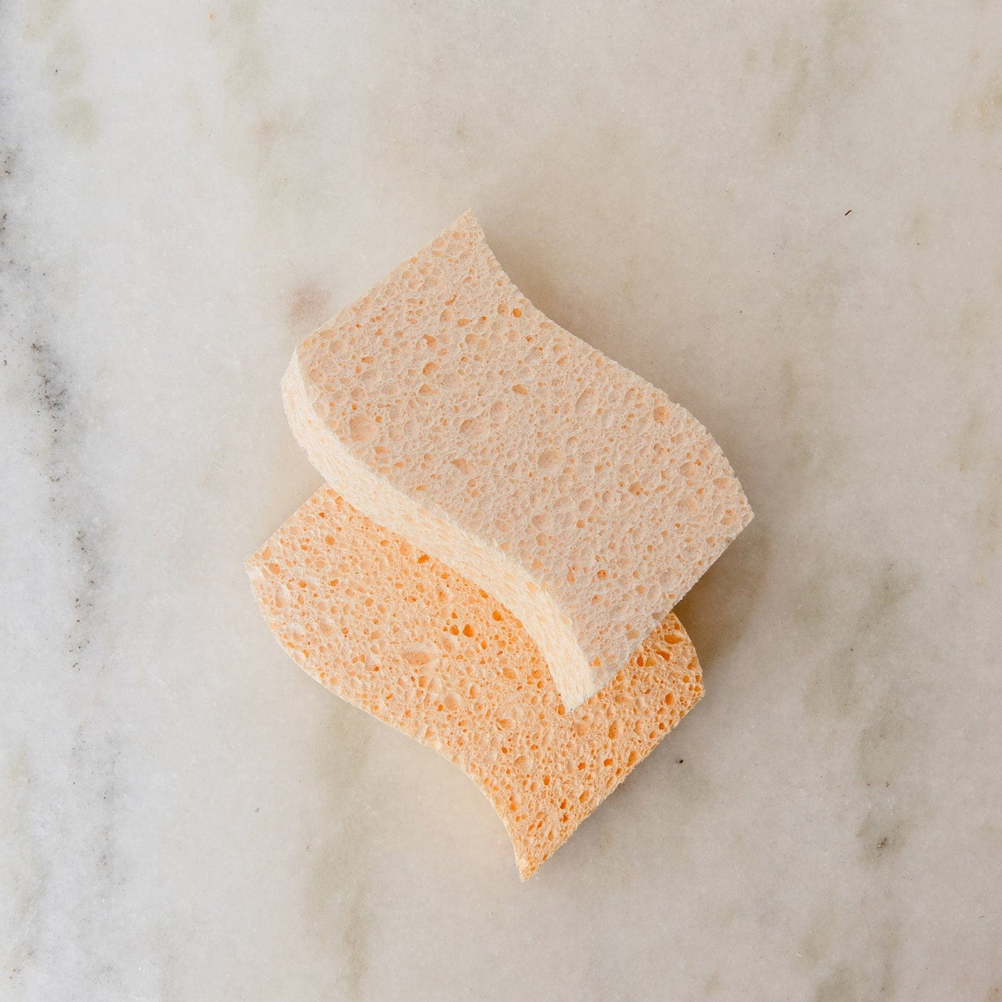 Two rectangular Cellulose Kitchen Sponges from ZeroWasteStore.com, one beige and one light orange, are stacked on a marble-like surface. These biodegradable sponges have a wavy edge and porous texture for effective cleaning.