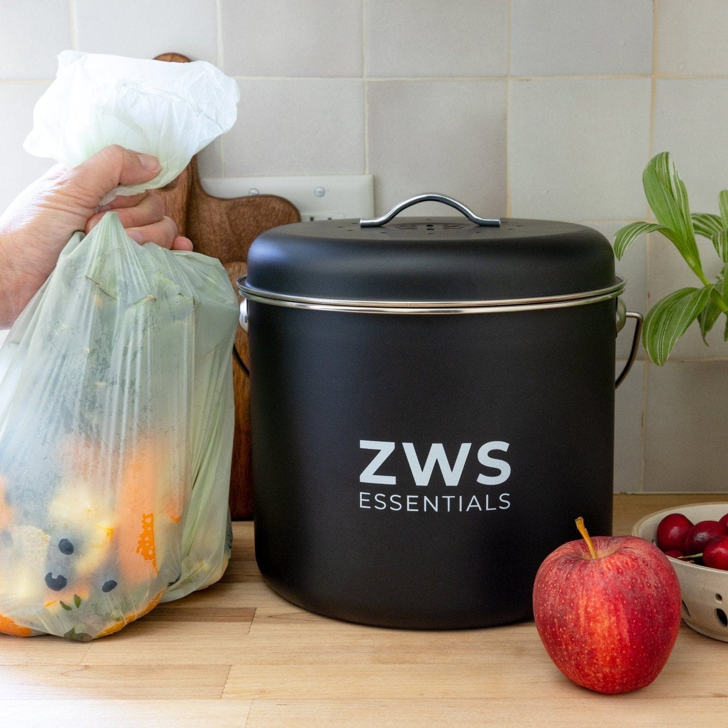 A hand holds a compostable bag with food scraps next to a black Countertop Compost Bin from ZeroWasteStore.com on the counter, surrounded by an apple, cherries, and a plant—ideal for any kitchen compost setup.