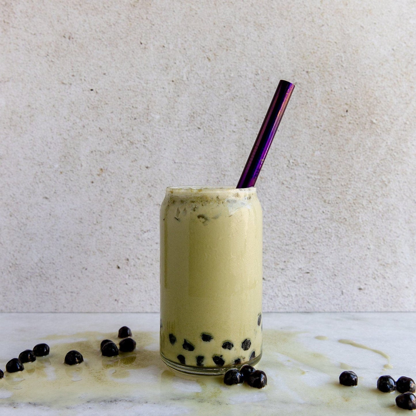 A glass of bubble tea with black tapioca pearls and a ZeroWasteStore.com Boba Style Stainless Steel Straw sits on a marble surface. Some pearls and spilled tea are scattered nearby, with a plain light background.