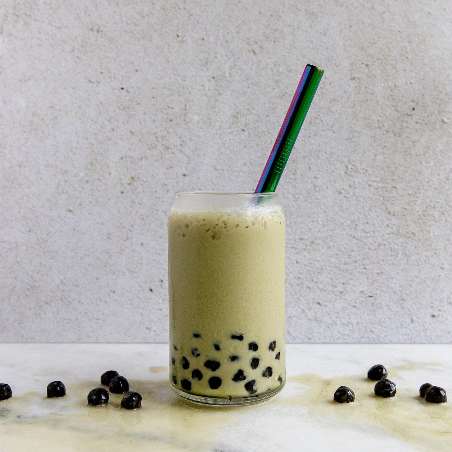Enjoy a refreshing bubble tea with black tapioca pearls, topped with foam, and sip sustainably using the ZeroWasteStore.com Stainless Steel Straw - Straight, Bent, or Boba Style. Some pearls are scattered on the marble surface for a stylish touch.