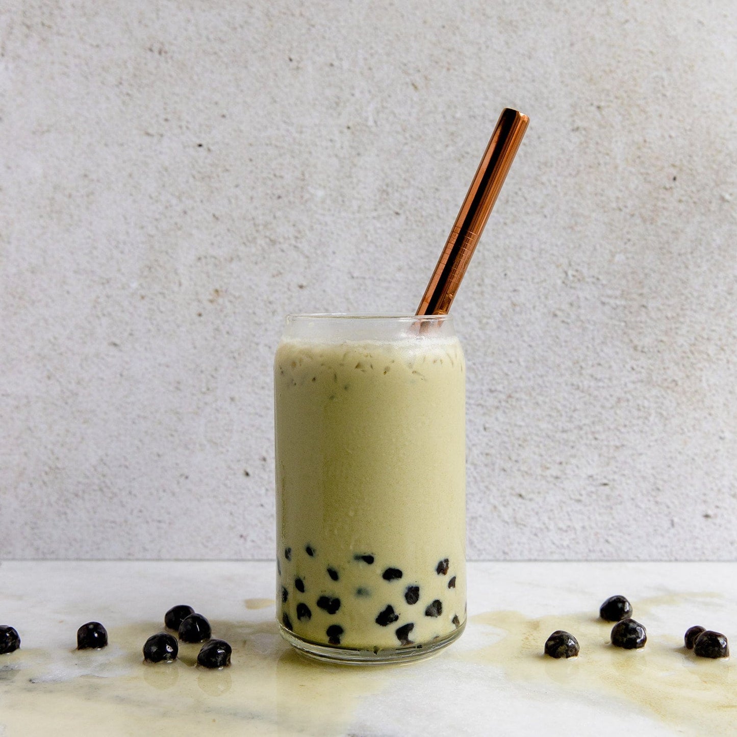 Enjoy a refreshing glass of bubble tea with black tapioca pearls and a light beige drink, sipped through the ZeroWasteStore.com Stainless Steel Straw—choose straight, bent, or boba style—for a plastic-free treat. Pearls scatter on marble for an eco-friendly touch.