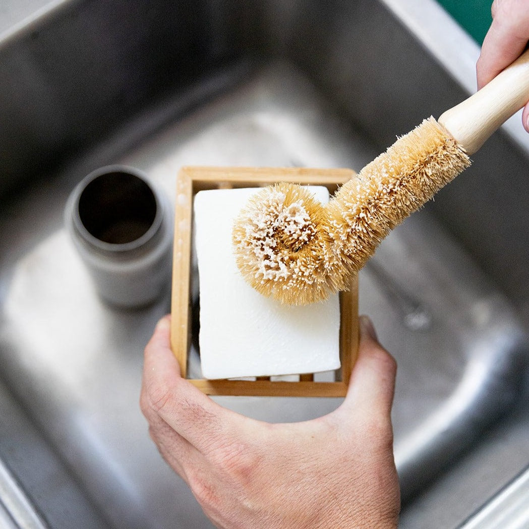 A hand uses the ZeroWasteStore.com Bottle Cleaning Brush to scrub a white bar of soap in a wooden holder over a kitchen sink, with a metal container inside the sink.