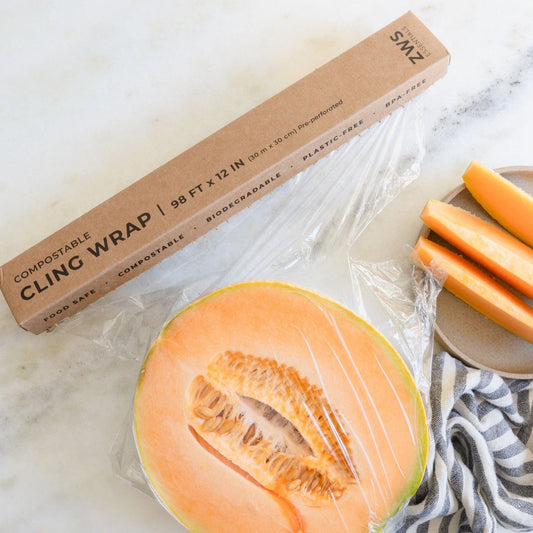 A halved cantaloupe wrapped in ZeroWasteStore.com Compostable Cling Wrap rests on a marble surface beside its box and a plate of cantaloupe slices.