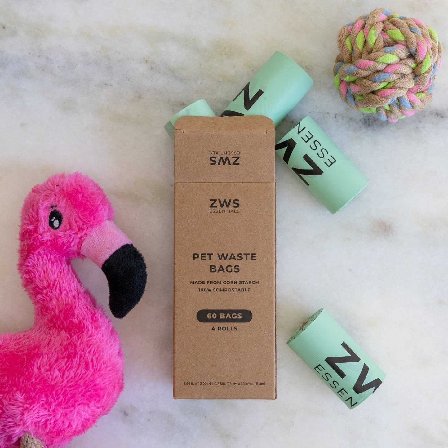 A box of ZeroWasteStore.com Compostable Pet Waste Bags (three green rolls), a pink flamingo plush toy, and a colorful rope ball are displayed on a white surface.