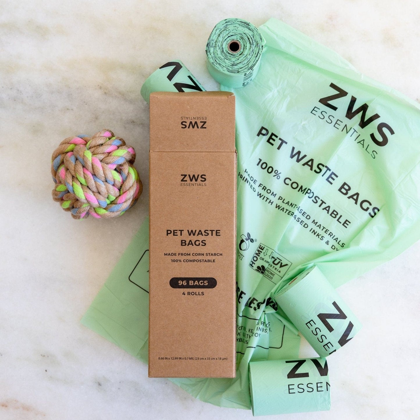 A box of ZeroWasteStore.com Compostable Pet Waste Bags, several green rolls, an unfolded bag, and a colorful rope toy are arranged on a white marble surface.
