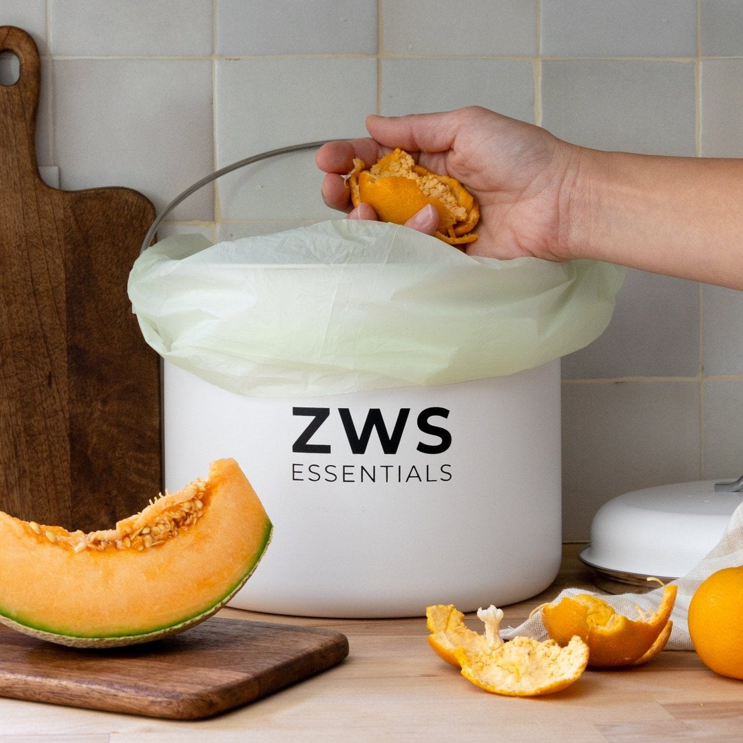 A hand drops fruit scraps into a white Countertop Compost Bin from ZeroWasteStore.com on a kitchen counter, surrounded by a cutting board, melon slice, and orange peels.