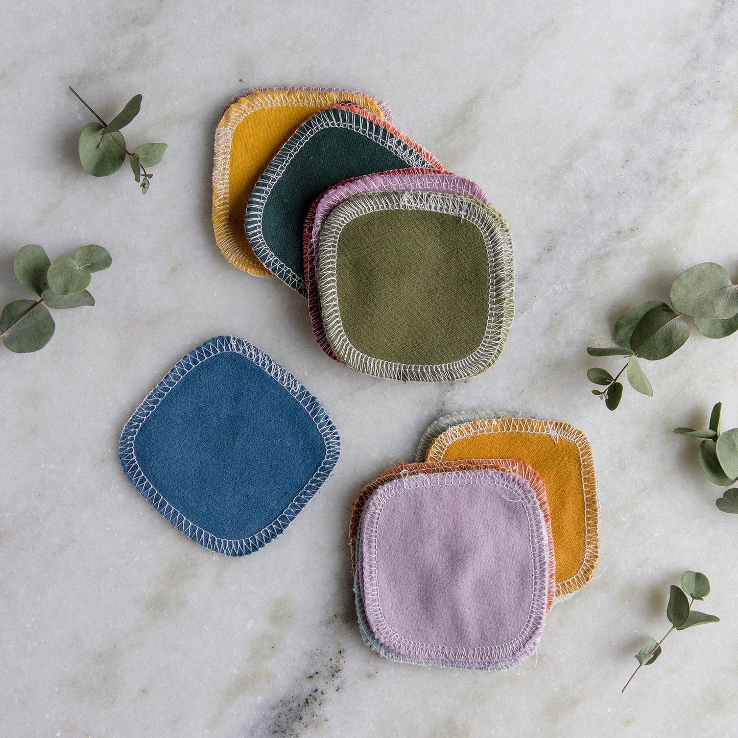 ZeroWasteStore.com Reusable Cotton Rounds in blue, pink, green, yellow, and mustard have stitched edges and a square shape. They're shown on a marble surface with eucalyptus sprigs for a colorful, eco-friendly skincare option.