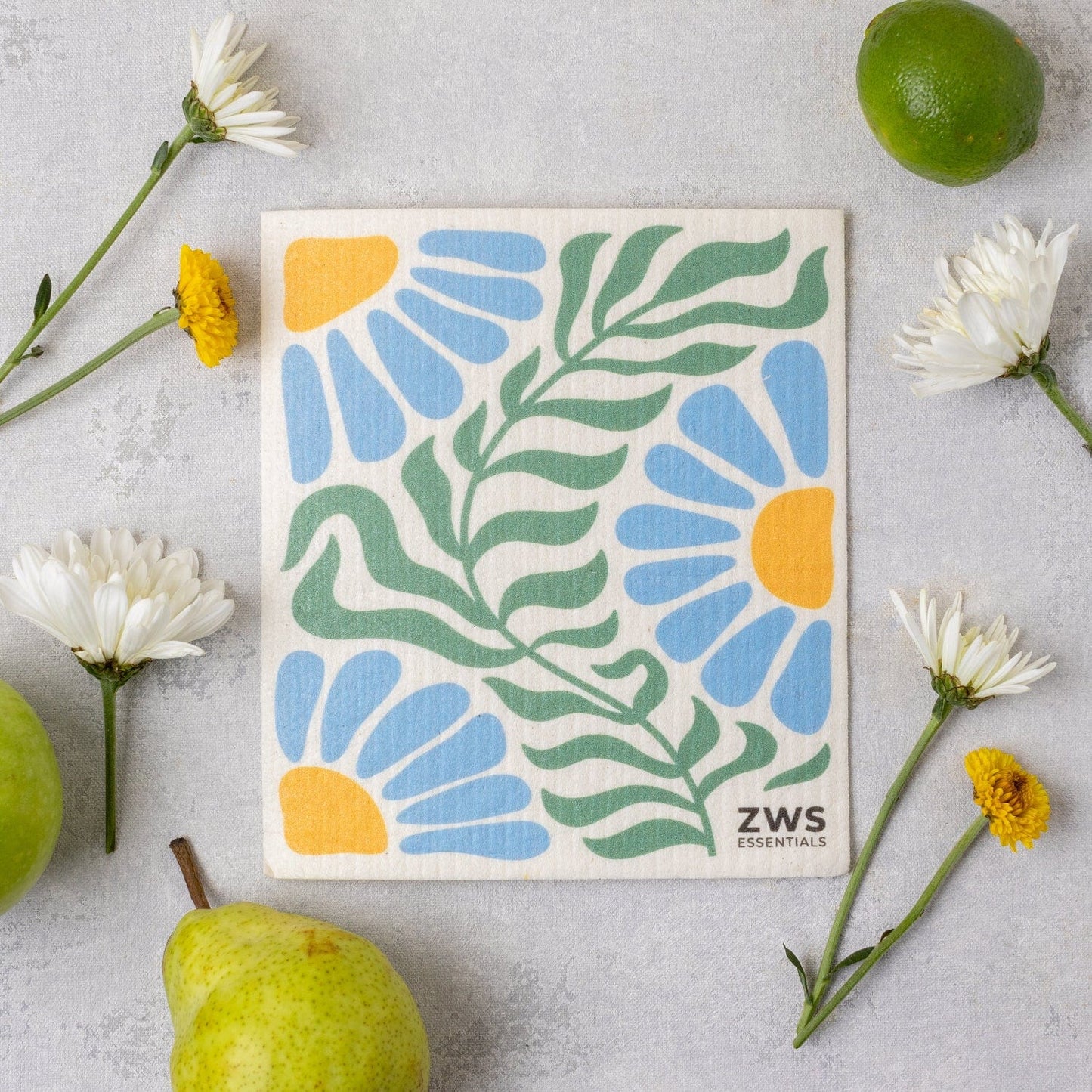A square Swedish Dish Cloth from ZeroWasteStore.com with a blue, yellow, and green floral design lies on a gray surface amid white flowers, a yellow flower, lime, and pear—an eco-friendly paper towel alternative.