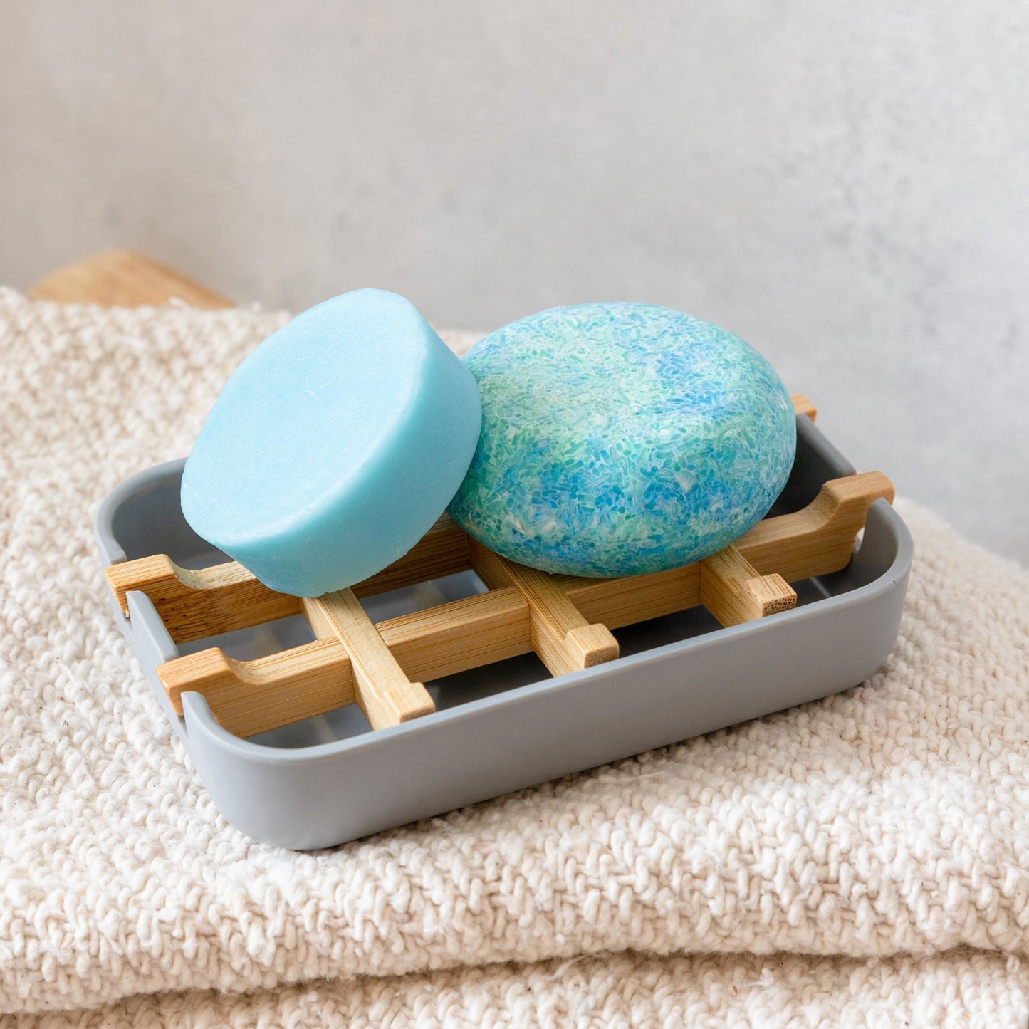 Two round blue soap bars sit on the ZeroWasteStore.com Bamboo Soap Dish inside a gray tray, all placed atop a folded beige textured towel.