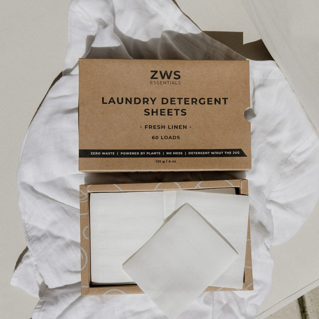 A box of ZeroWasteStore.com Laundry Detergent Sheets rests on white fabric. The open box displays white sheets, with packaging featuring Fresh Linen scent, 60 loads per pack, zero waste, and plant-based ingredients.