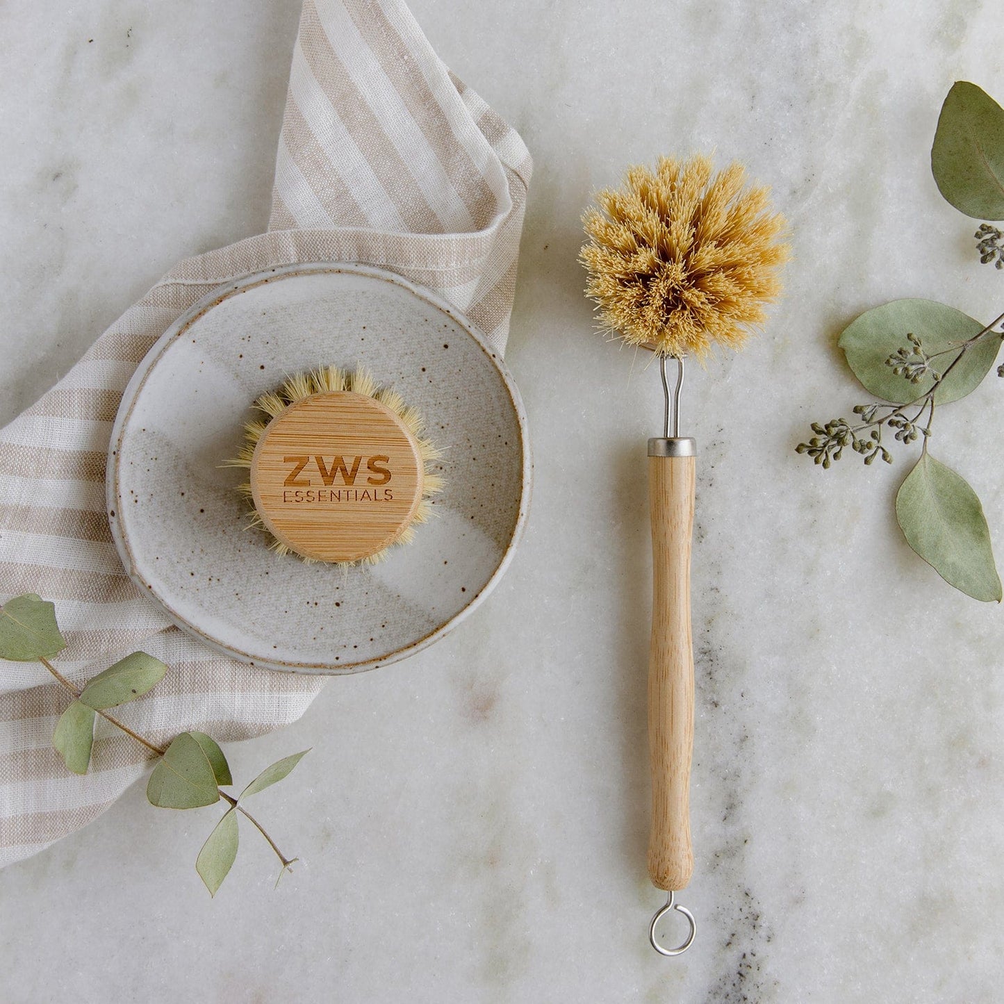 The Long Handled Dish Brush Mini Kit by ZeroWasteStore.com, featuring two natural bristle brushes with bamboo handles, rests on a marble surface near a striped cloth, small plate, and green eucalyptus leaves.