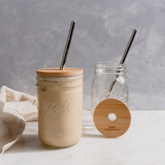 Two mason jars from the ZeroWasteStore.com Mason Jar Kit—featuring Bamboo Lids and Stainless Steel Straws—sit on a light surface. One jar contains iced coffee, while the other is empty with its lid and straw beside it; a beige cloth is on the left.