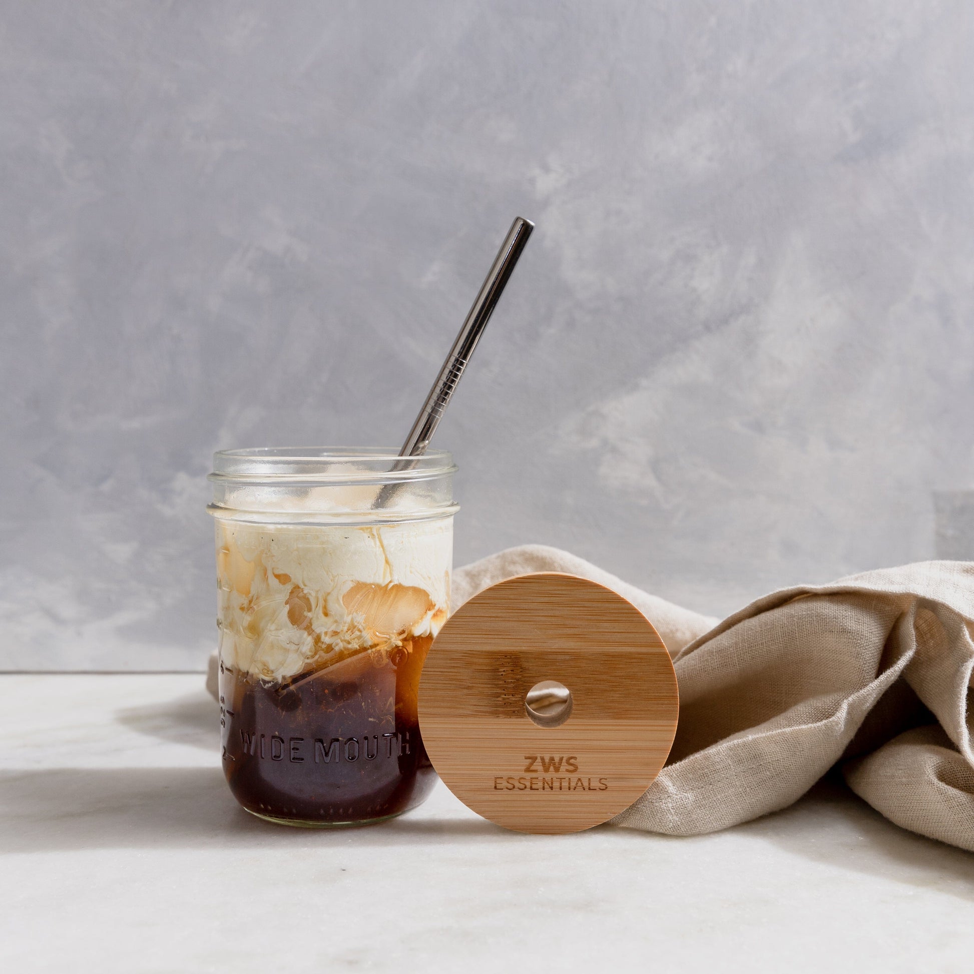 A Mason Jar Kit from ZeroWasteStore.com—with a glass mason jar, bamboo lid, and stainless steel straw—holds iced coffee and cream, while the branded lid rests on a beige cloth against a light gray background.