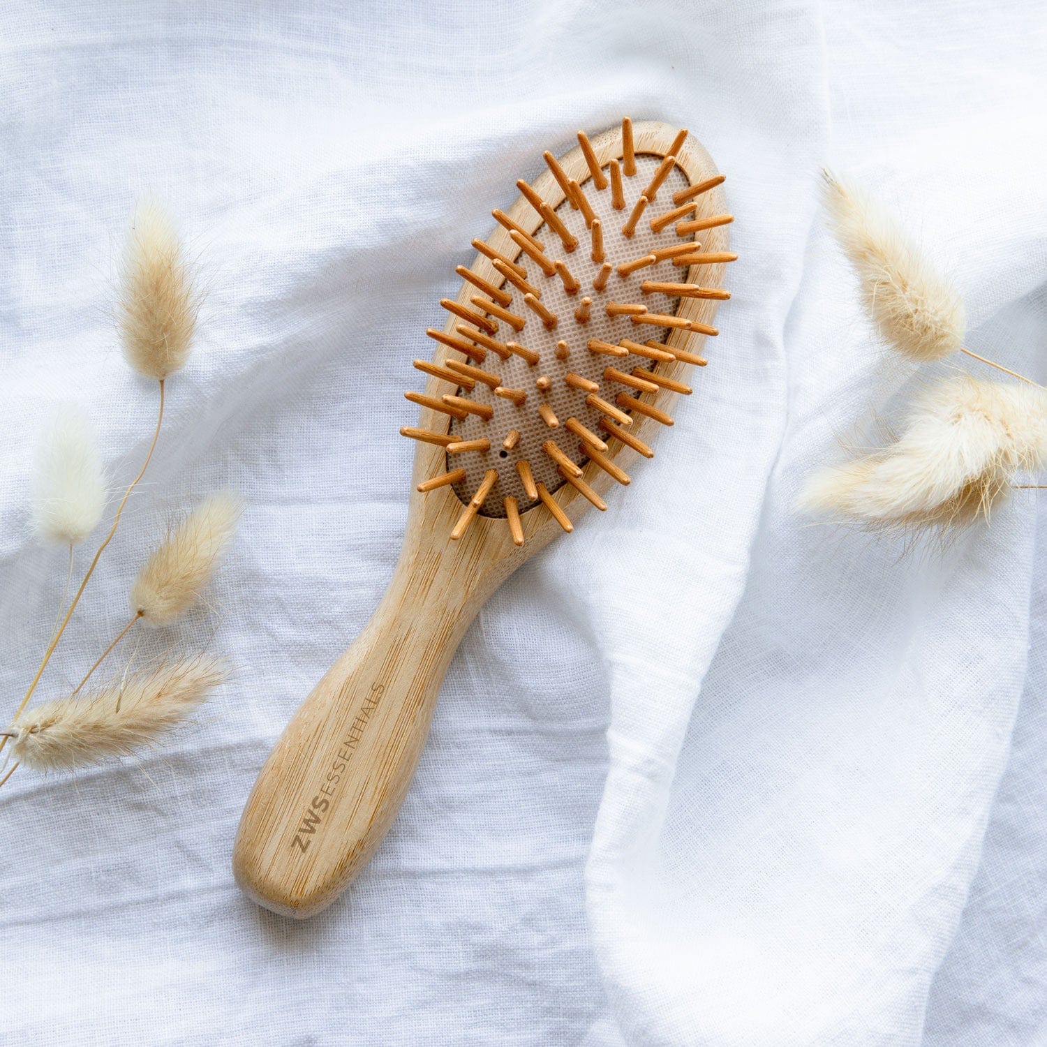 The Mini Bamboo Hair Brush by ZeroWasteStore.com rests on a white fabric with dried grasses—this 100% bamboo, plastic-free, compostable brush is the perfect zero waste choice for eco-conscious hair styling.