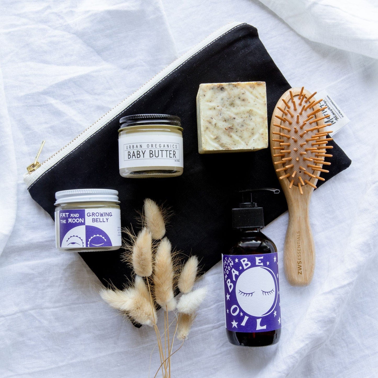 A flat lay of baby care items on white fabric: a black zip pouch, a ZeroWasteStore.com Mini Bamboo Hair Brush (plastic free, 100% bamboo), a bar of soap, a Babe Oil bottle, Grooming Belly balm jar, and Baby Butter tin with dried grasses.