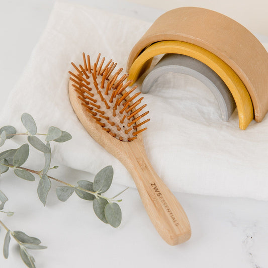 The Mini Bamboo Hair Brush by ZeroWasteStore.com, a 100% bamboo, plastic-free, compostable hair brush with brown bristles, rests on a white cloth beside a eucalyptus branch and a tan-yellow-gray wooden rainbow toy.